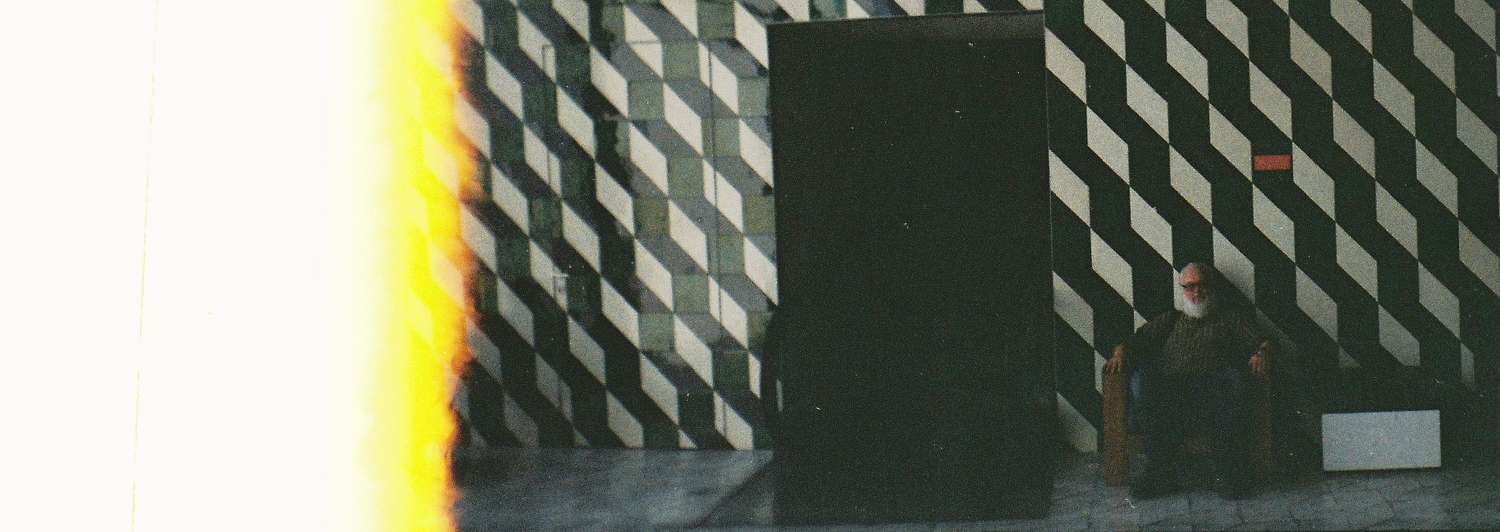 profile image - a bearded man is sitting on a chair against a geometric patterned wall with a dark passageway beside them and a light leak effect on the left side of the photo